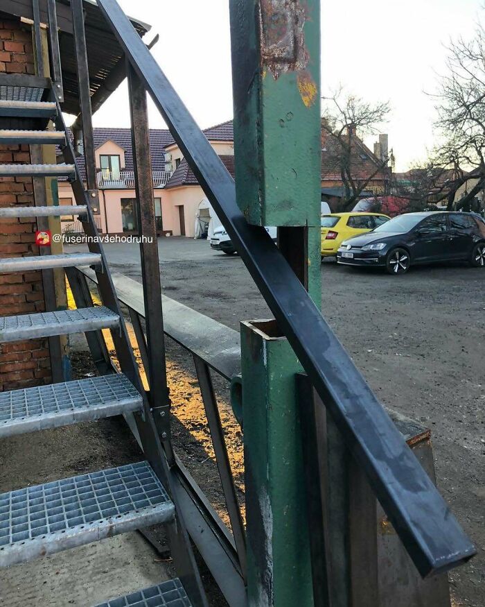 Metal construction design with a steel handrail awkwardly inserted into a green metal post outside near stairs and parked cars.