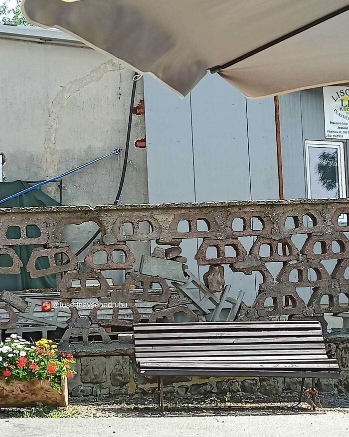 Rusty construction wall with irregular holes behind a broken bench and flowers, showcasing unusual design and construction.