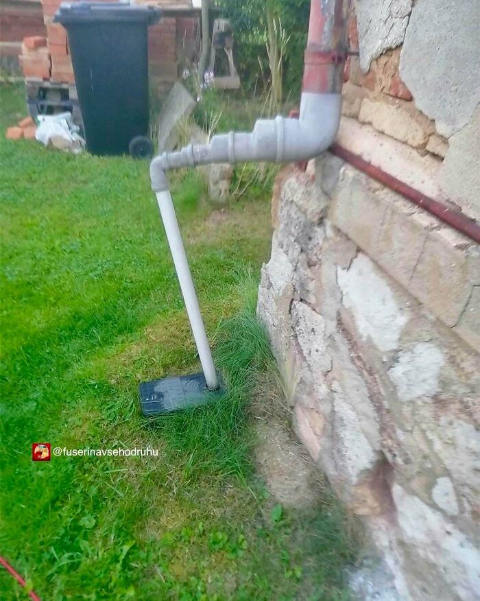 Unconventional construction design with a bent pipe draining into a closed container on grass near a brick wall.