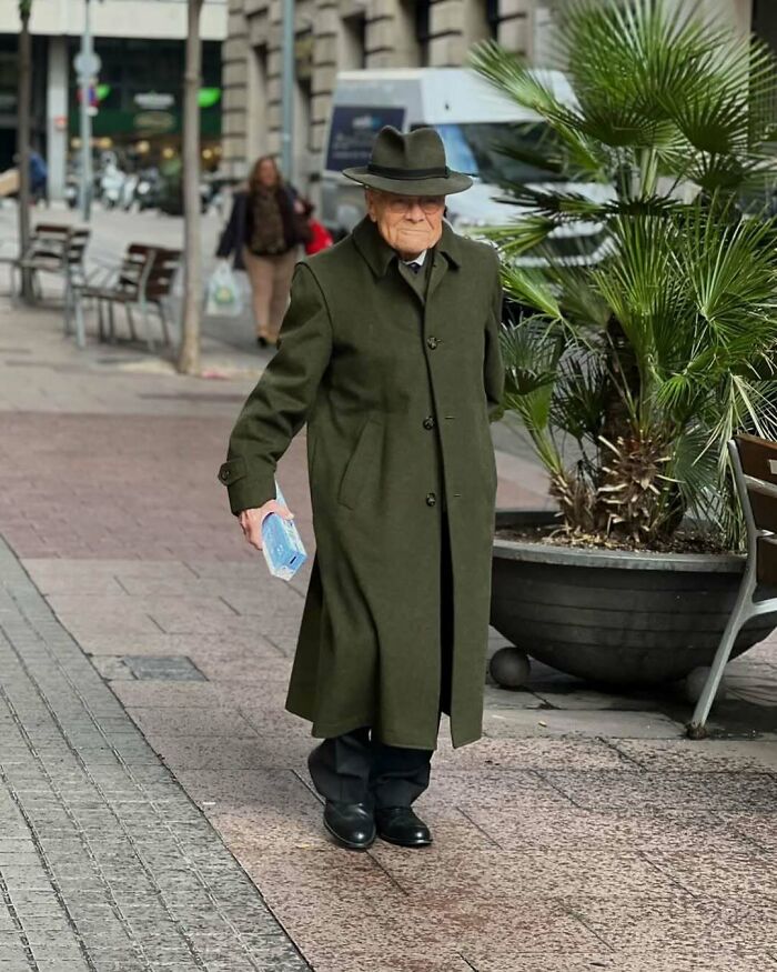 Stylish grandparent wearing a long green coat and hat walking confidently on a city sidewalk holding a face mask.