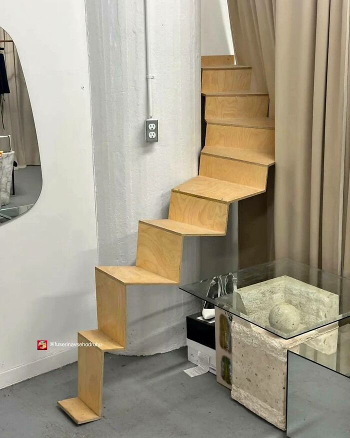 Zigzag wooden stairs with a unique design attached to a wall next to a glass table and beige curtain in a room.