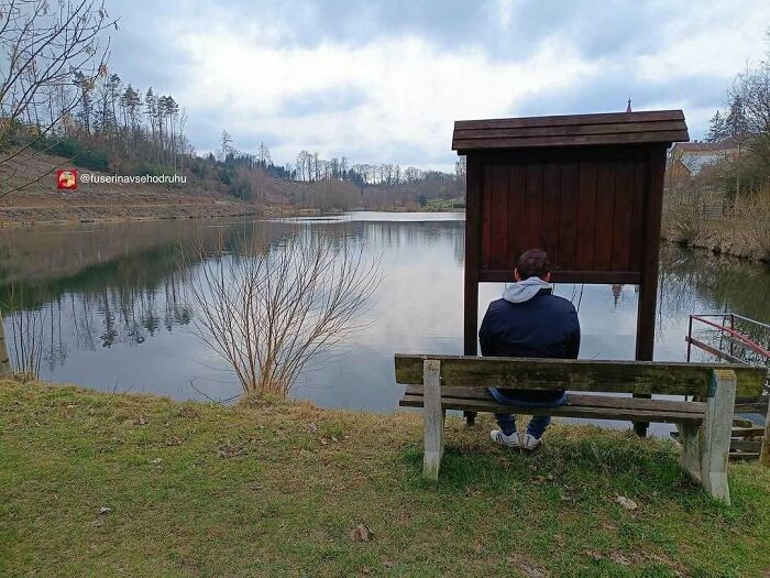 Man sitting on a bench facing a wooden construction with a roof by a calm lake in a quiet outdoor setting.