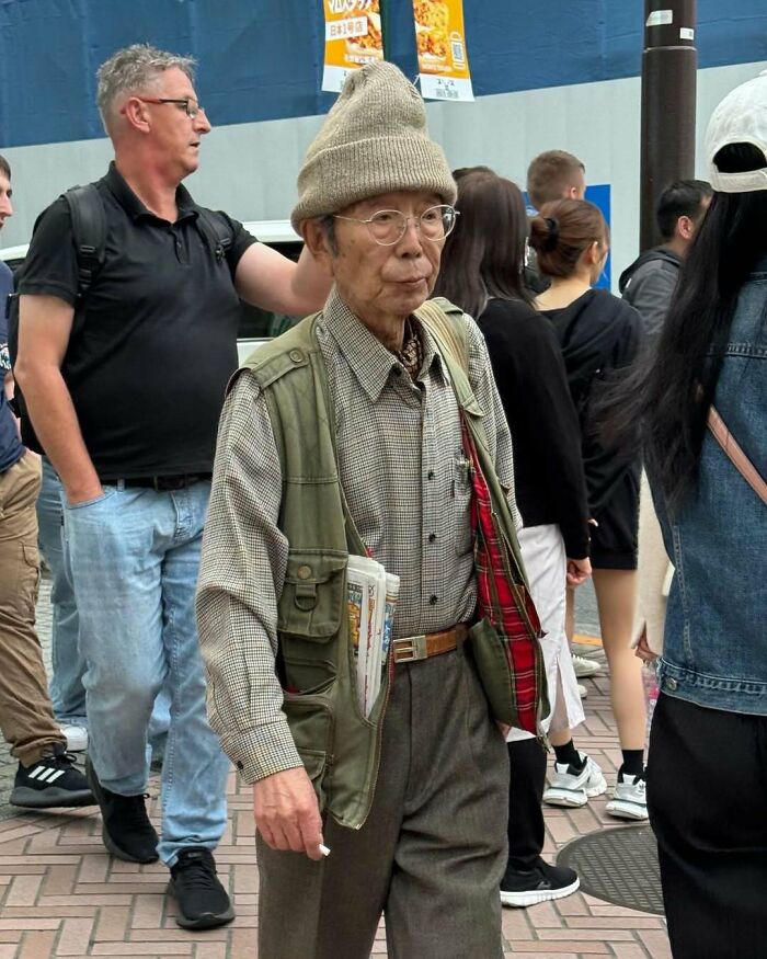 Stylish grandparent wearing a checkered shirt, green vest, and beige knit hat walking confidently in a busy urban setting.