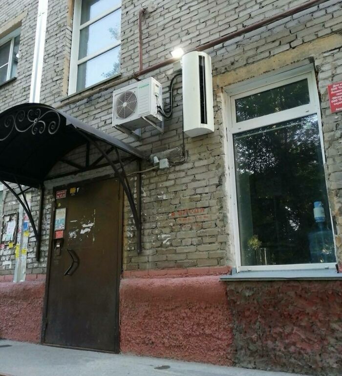 Air conditioning unit installed awkwardly above a door on a brick building, showing poor construction and design choices.