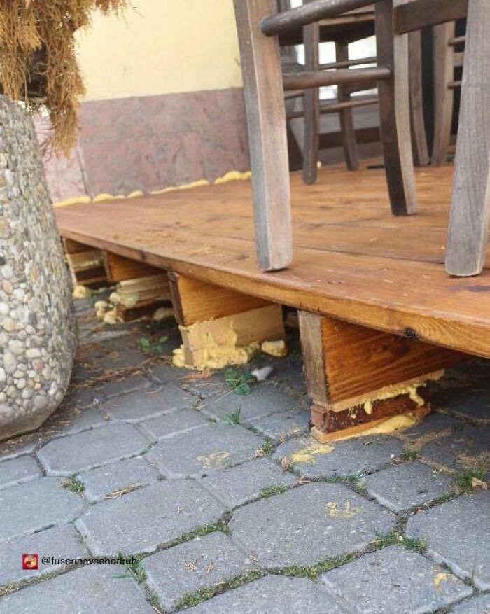 Wooden deck supported by uneven wooden blocks and expanding foam on a stone-paved outdoor area, highlighting unusual construction design.