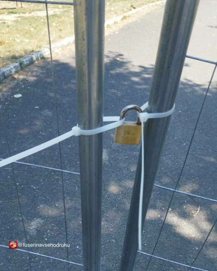 Construction design flaw showing a metal gate locked with a padlock secured by plastic zip ties on asphalt.