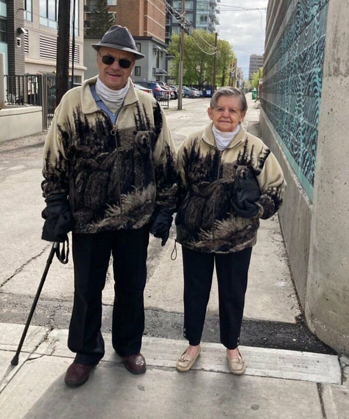 Elderly couple wearing matching stylish grandparent outfits with nature-themed jackets walking on a city sidewalk.