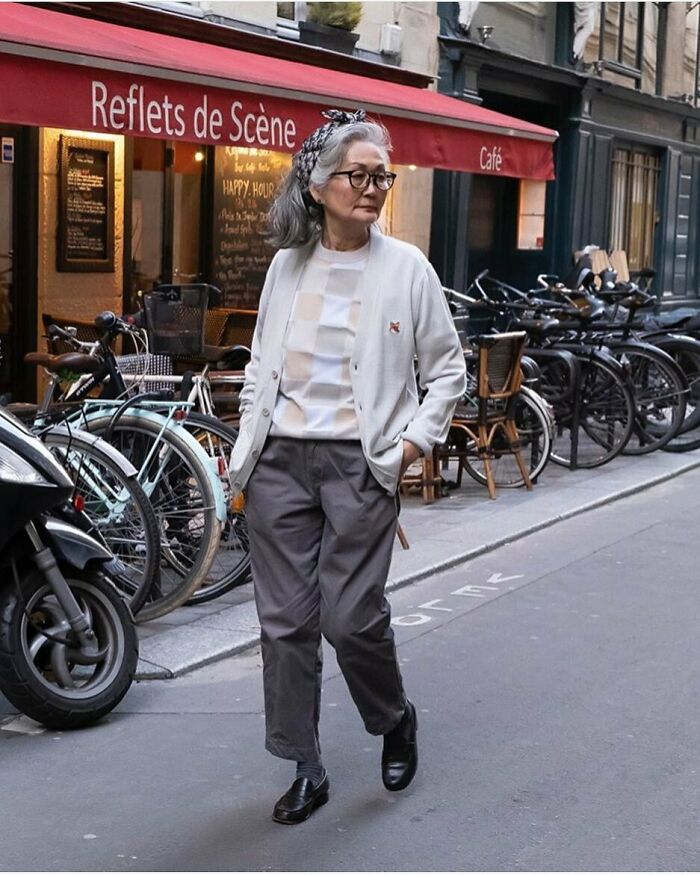 Stylish grandparent walking on city street wearing gray pants, white cardigan, and glasses with a headband in hair.