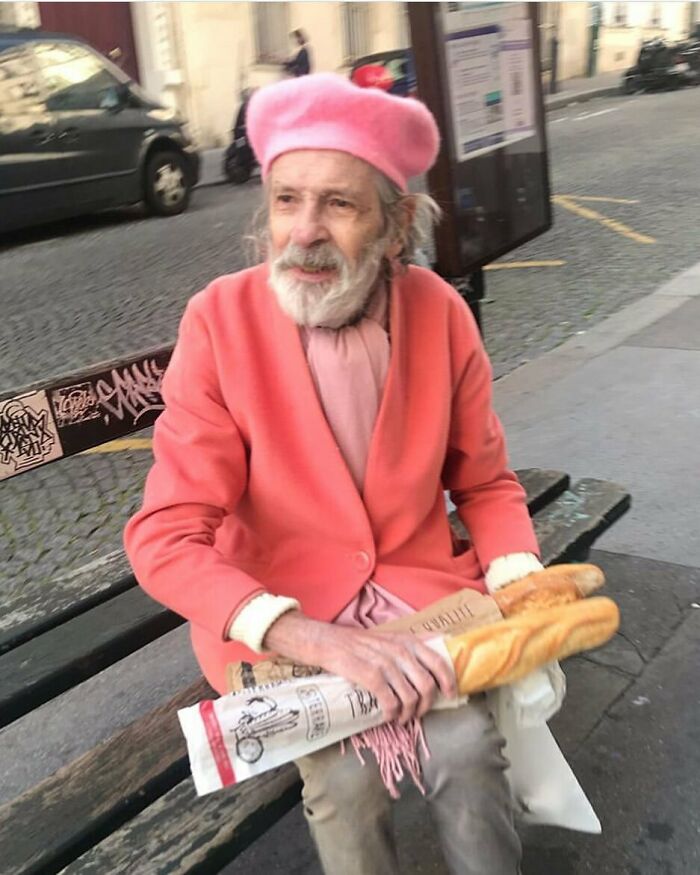 Elderly man in a stylish pink beret and coral coat sitting on a bench holding baguettes, showcasing cool and stylish grandparents fashion.