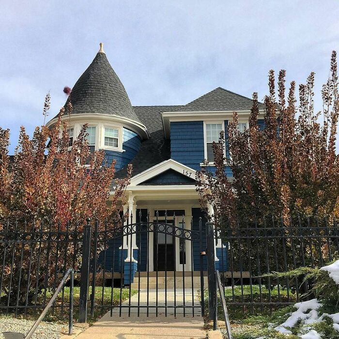 Blue old home with a turret and front porch behind a black iron fence surrounded by autumn trees and snow patches.