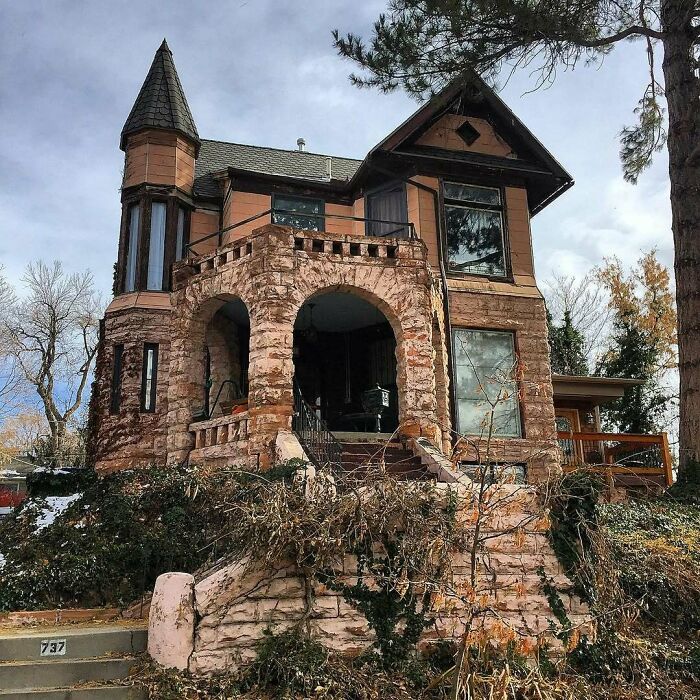 Stone old home with a turret and arched porch surrounded by trees and vegetation on a cloudy day.