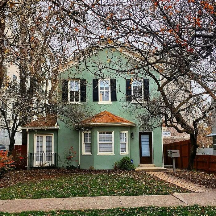 Charming adorable old home with green exterior and black shutters surrounded by leafless trees in a quiet neighborhood.