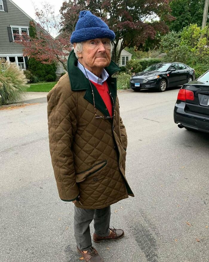 Stylish grandparent in a quilted jacket, red sweater, blue knit hat, and brown shoes standing on a suburban street.