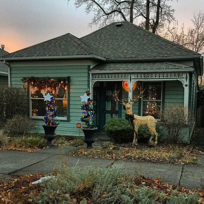 Cozy old home decorated with colorful lights and a glowing reindeer figure in the front yard at dusk.
