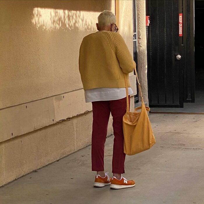 Stylish grandparent wearing a mustard sweater, red pants, and orange sneakers carrying a matching tote bag outdoors.