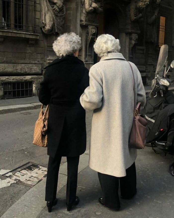 Two stylish grandparents wearing elegant coats and carrying handbags, walking on a city street in cool outfits.