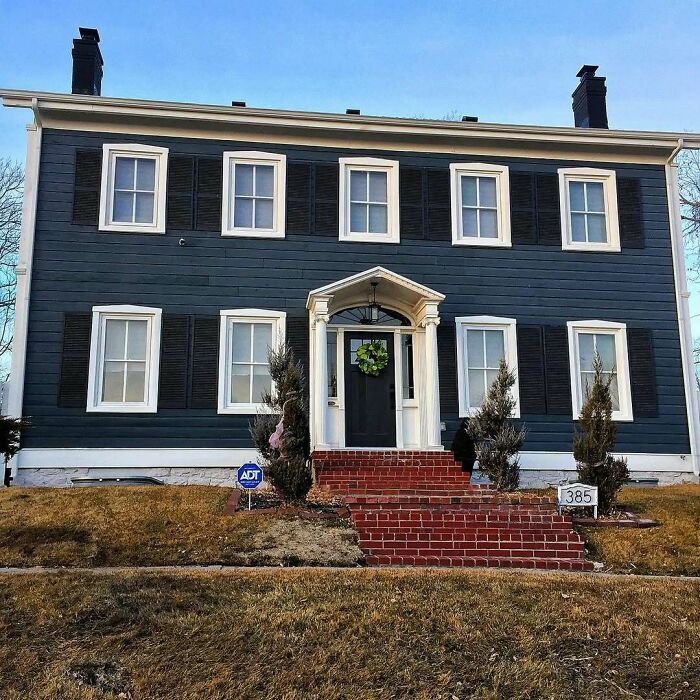Classic charming old home with dark siding and white trim, featuring a brick stairway and decorative greenery at the entrance.