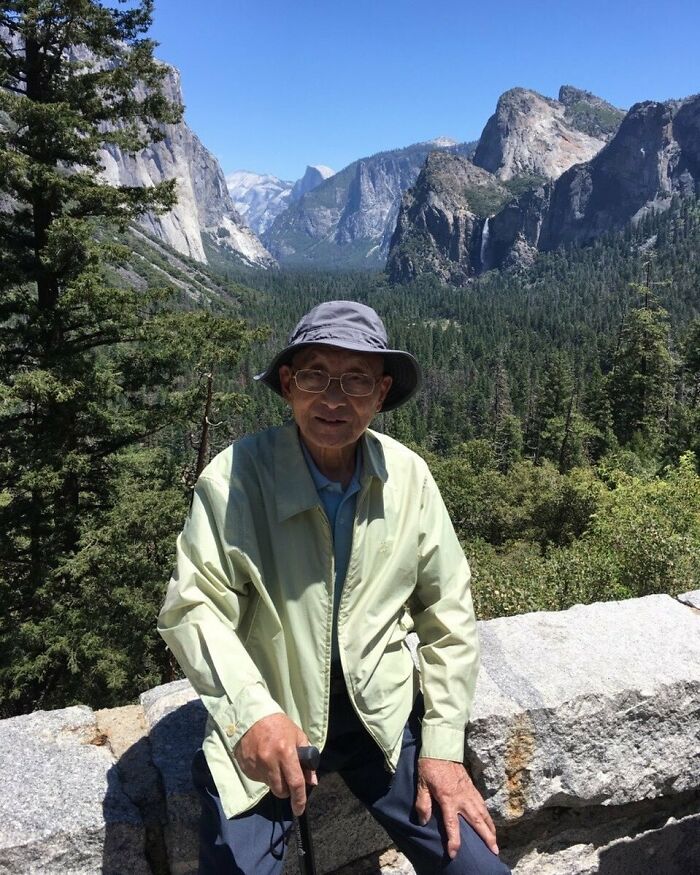 Elderly man wearing a light green jacket and hat, posing outdoors with mountains and forest in the background, stylish grandparents.