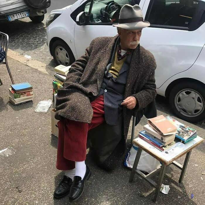 Stylish grandparent wearing a layered outfit with a hat, sitting outdoors near stacks of books by parked cars.
