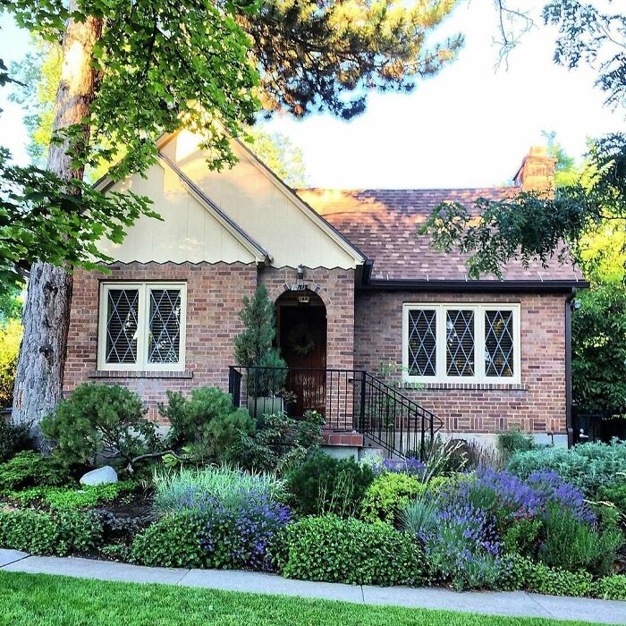 Charming old brick home with diamond-paned windows surrounded by lush greenery and colorful garden in a sunny neighborhood.