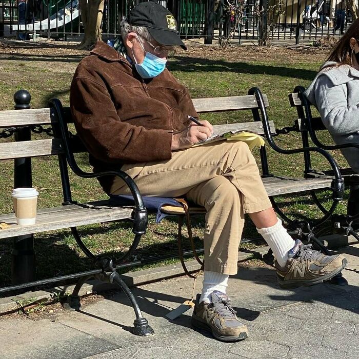 Elderly man wearing stylish casual outfit with brown jacket, beige pants, and sneakers, sitting on a park bench writing.