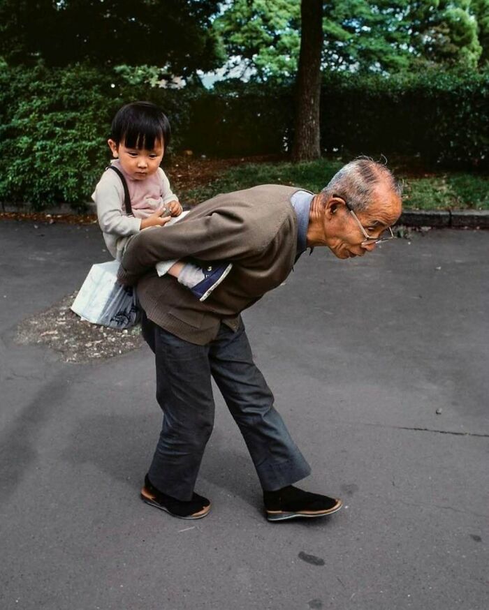 Elderly man in stylish casual outfit giving a piggyback ride to a child outdoors, showcasing cool grandparents' fashion.