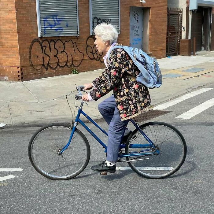 Stylish grandparent riding a blue bicycle wearing a floral jacket and light blue backpack on an urban street.