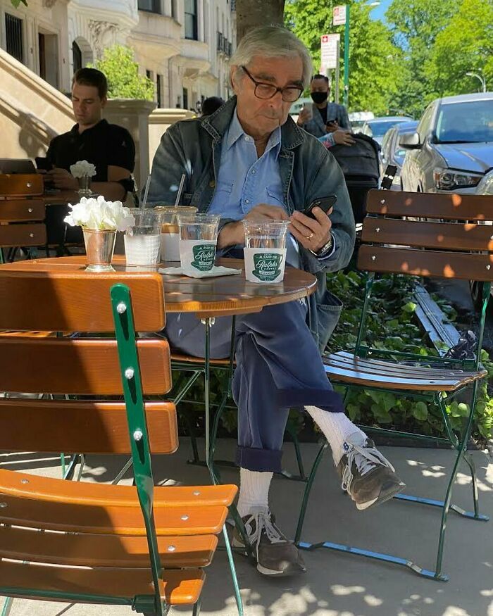 Older man in stylish denim jacket and glasses sitting outdoors at a cafe with coffee cups, showing cool and stylish grandparents fashion.
