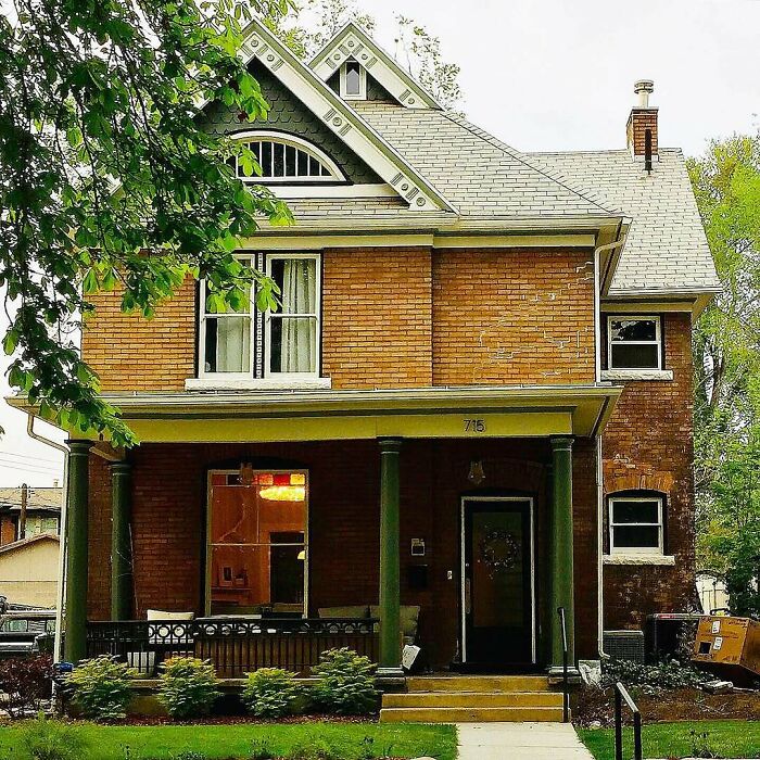 Brick old home with green pillars and a porch surrounded by trees and shrubs in a residential neighborhood.