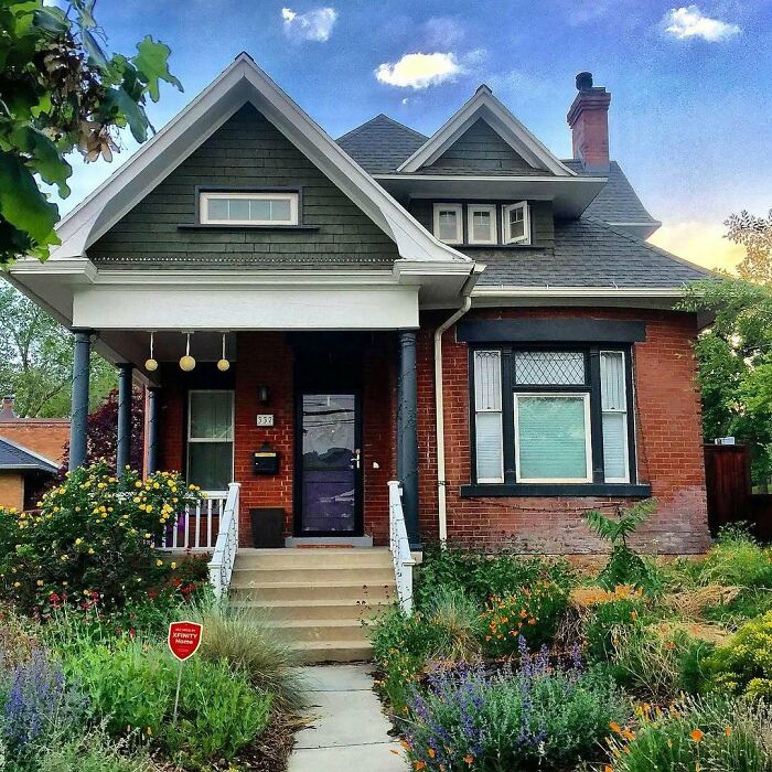 Adorable and lovely old home with brick exterior, green gable roof, and lush front garden under a partly cloudy sky.