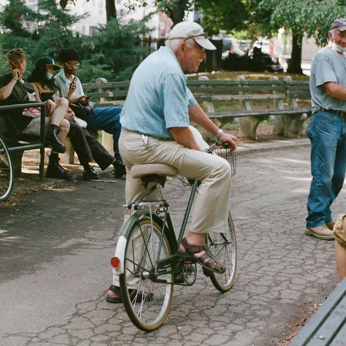 Stylish grandparent wearing light blue shirt and beige pants riding a bicycle in a park with others nearby.