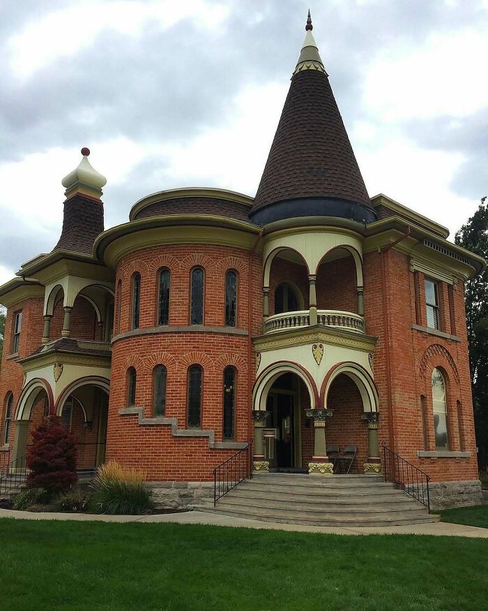 Victorian-style old home with rounded tower and arched windows surrounded by green lawn under cloudy sky.