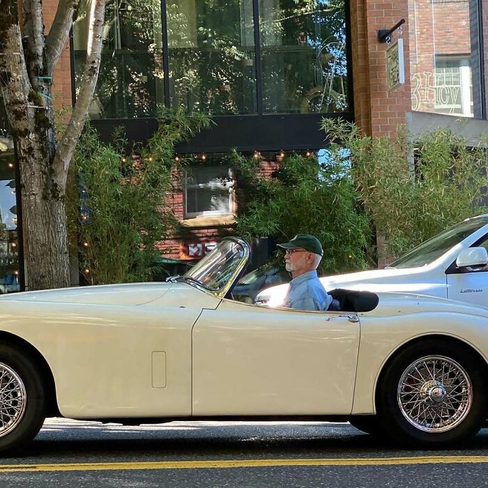 Stylish grandparent wearing a cap and glasses driving a vintage convertible car on a sunny urban street.