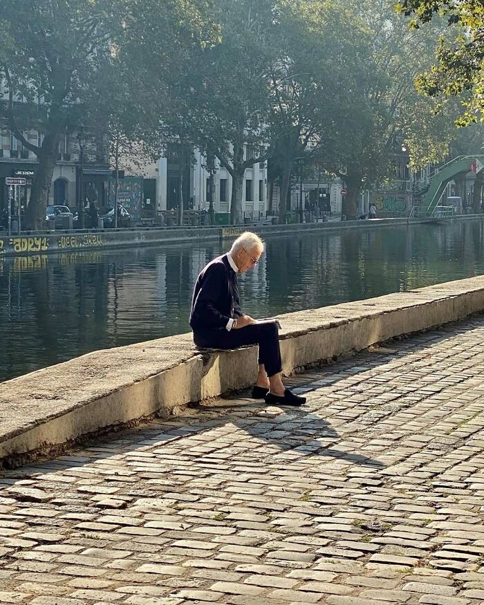 Elderly man in stylish outfit sitting by a canal, reading, showcasing cool and stylish grandparents fashion.