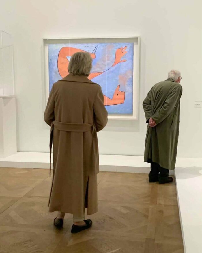 Two stylish grandparents wearing long coats, admiring modern art in a bright gallery with wooden floors.