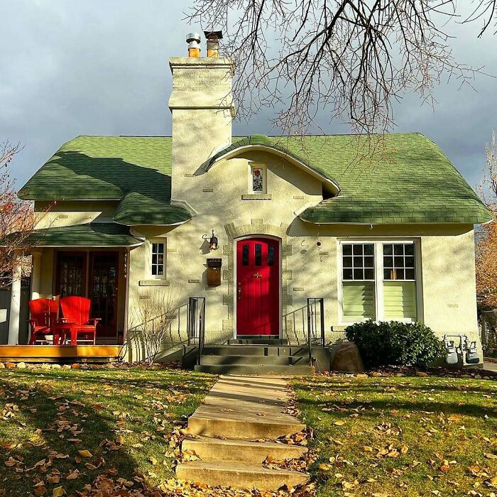 Charming old home with green roof, red door, front porch seating, and a pathway surrounded by autumn leaves in a cozy neighborhood.