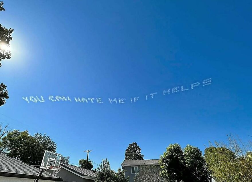 Clear blue sky with fluffy cloud letters forming a message above a quiet suburban neighborhood scene.
