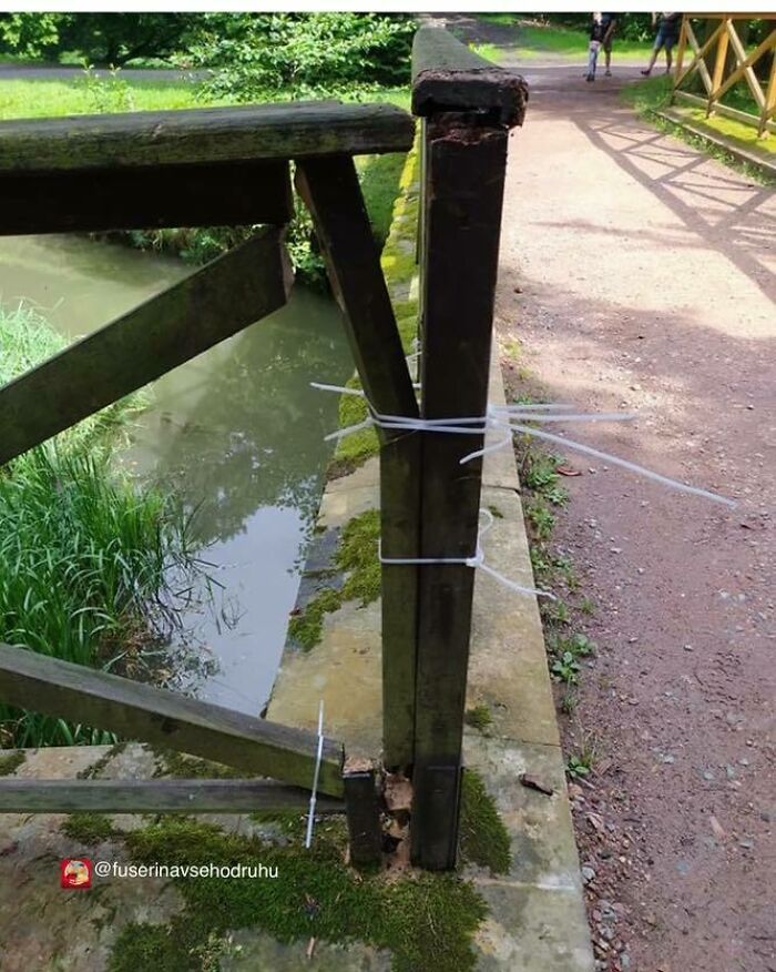 Damaged wooden fence post poorly repaired with zip ties on a pathway by a small body of water, illustrating flawed construction design.