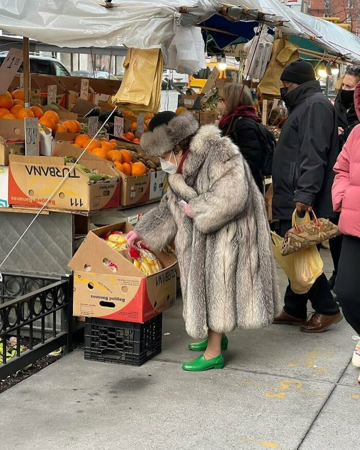 Stylish grandparent wearing a fur coat and green shoes shopping for fruit at an outdoor market.