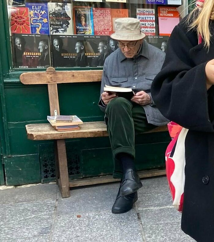 Stylish grandparent sitting on a bench reading a book wearing a bucket hat, gray jacket, green pants, and black shoes.