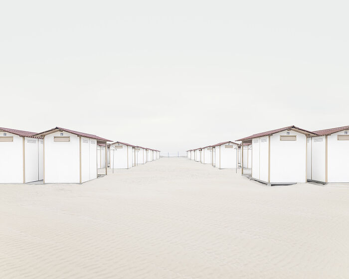 Rows of simple white beach cabins with red roofs on a sandy shore, showcasing minimalism in architecture and design.