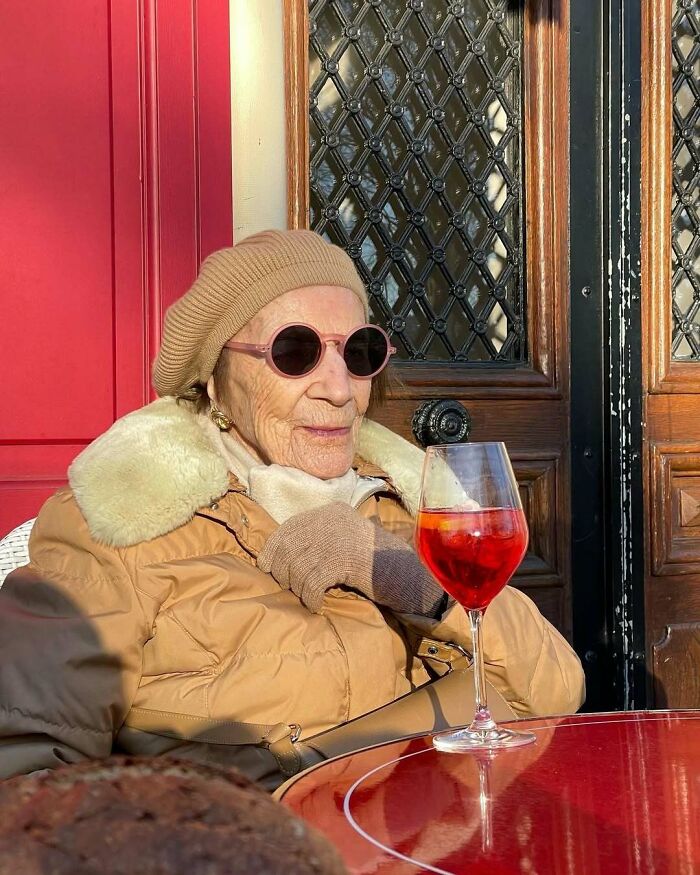 Stylish grandparent in beige beret, sunglasses, and coat enjoying a drink outdoors against a red and wooden background.