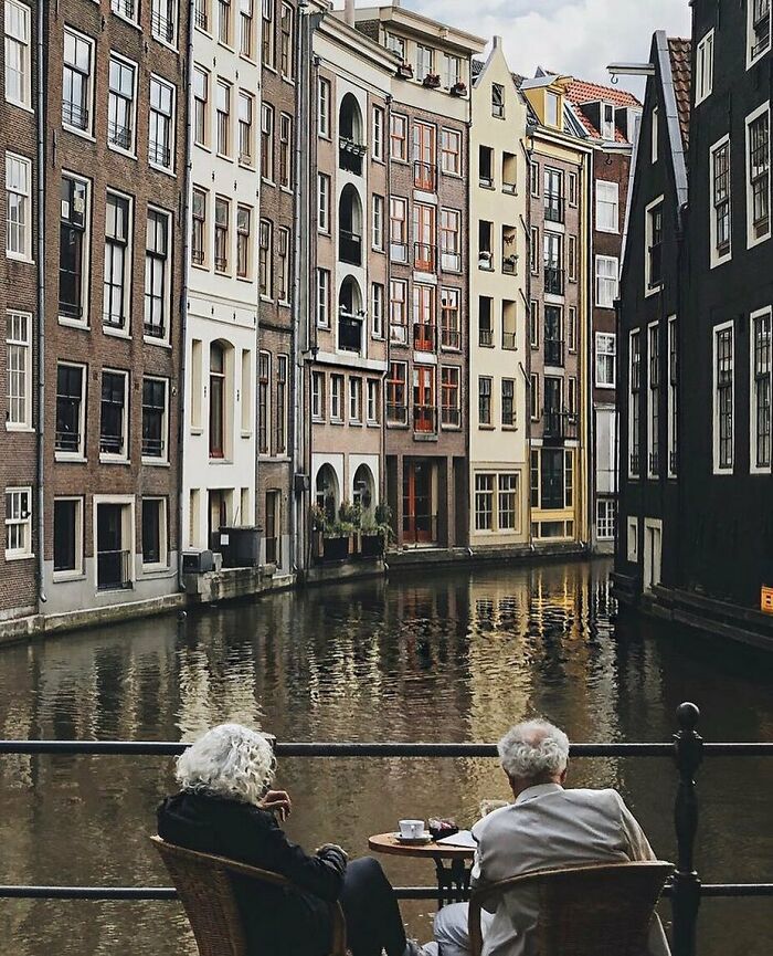 Two stylish grandparents enjoying coffee by a canal with reflections of tall, charming European buildings in the water.