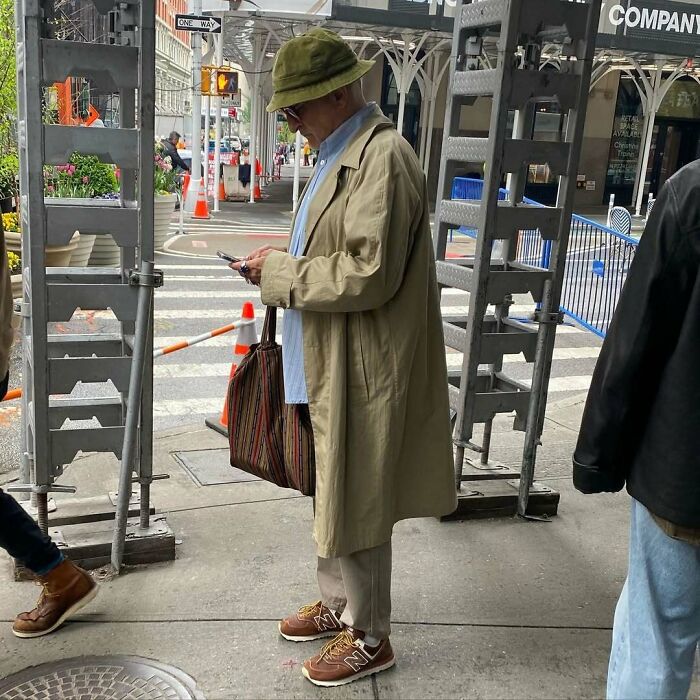 Stylish grandparent wearing a trench coat and bucket hat, standing on a city sidewalk with a striped bag.