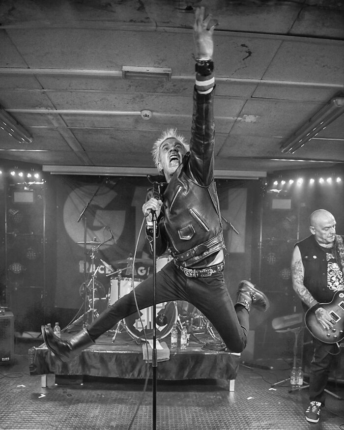 Black-and-white photo of a dynamic punk rock singer mid-jump on stage during a live music performance.