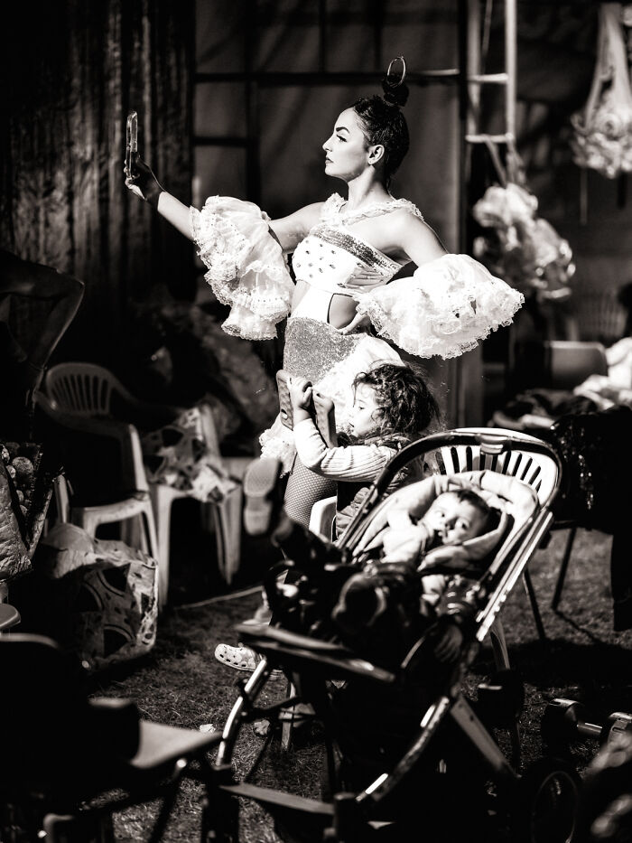 Black-and-white photo of a dancer in costume taking a selfie while children play nearby in a candid moment.