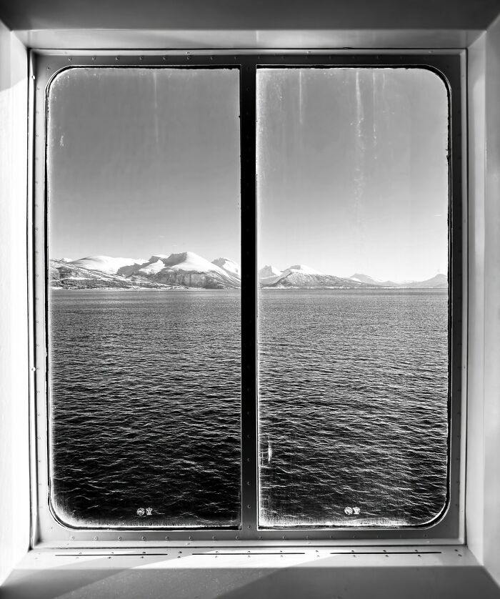 Black-and-white photo of a snowy mountain landscape viewed through a window over a calm sea.