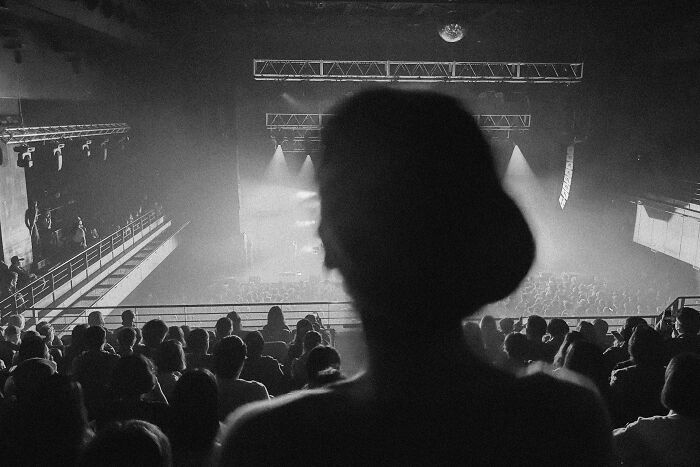 Black-and-white photo showing a large audience in a theater or concert hall from behind a silhouetted figure.