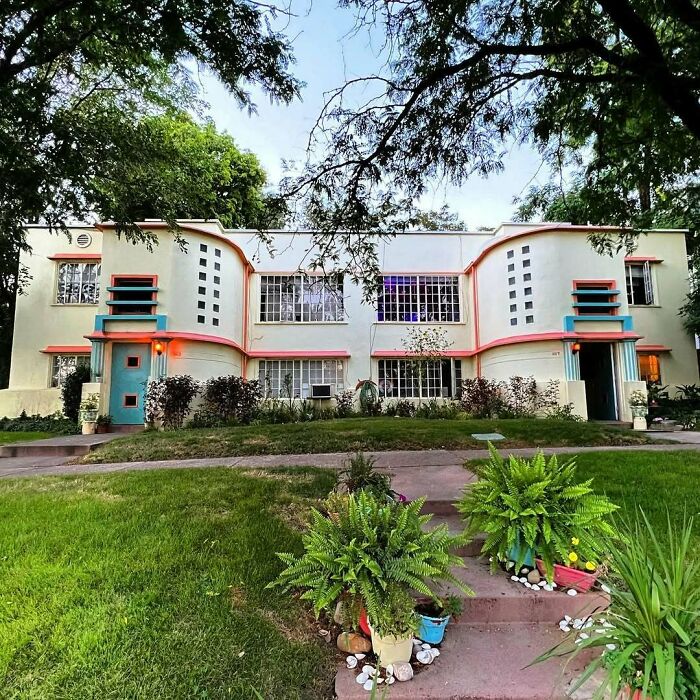 Art deco style old home with pastel colors, large windows, and lush green lawn surrounded by trees and plants.