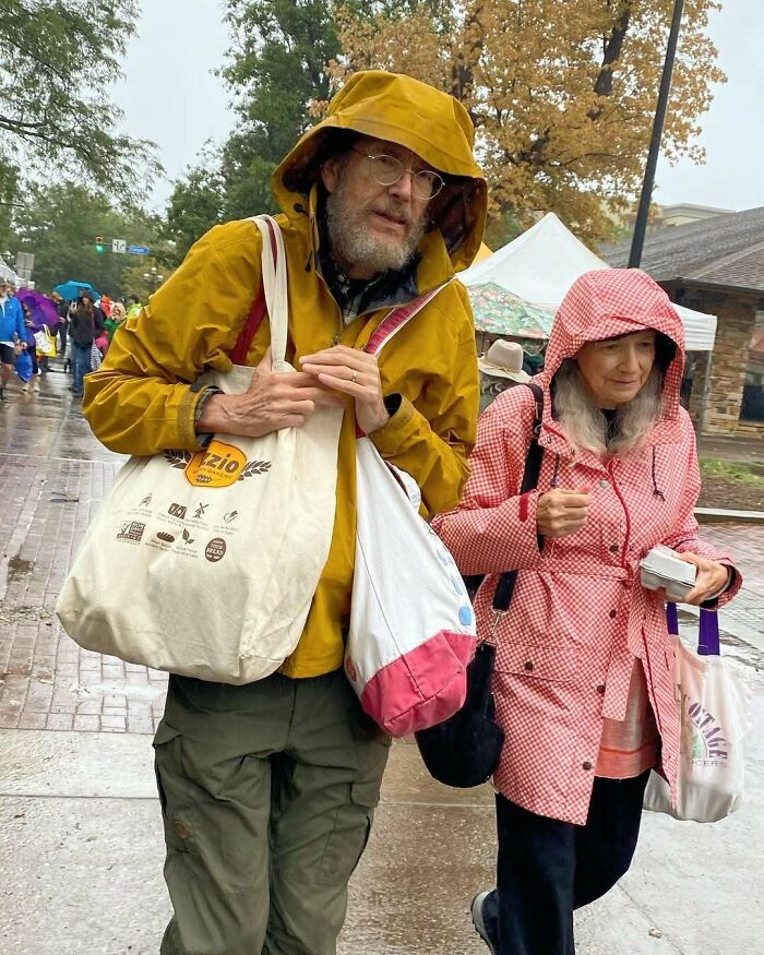 Stylish grandparents in colorful raincoats carrying bags while walking at an outdoor market on a rainy day.